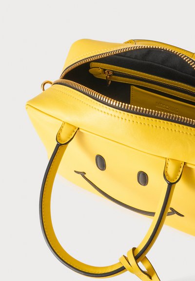 Sac à main en cuir jaune avec des détails de smiley noir, partiellement dézippé, montrant l'intérieur noir et une poche intérieure zippée.