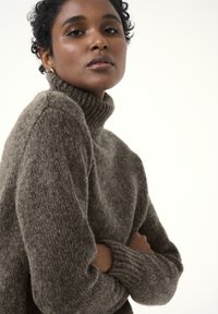Hochkragen, grauer Strickpullover mit weicher Textur. Mit gerippten Ärmelbündchen und lockerer Passform. Ein Model posiert mit verschränkten Armen und präsentiert das Kleidungsstück.