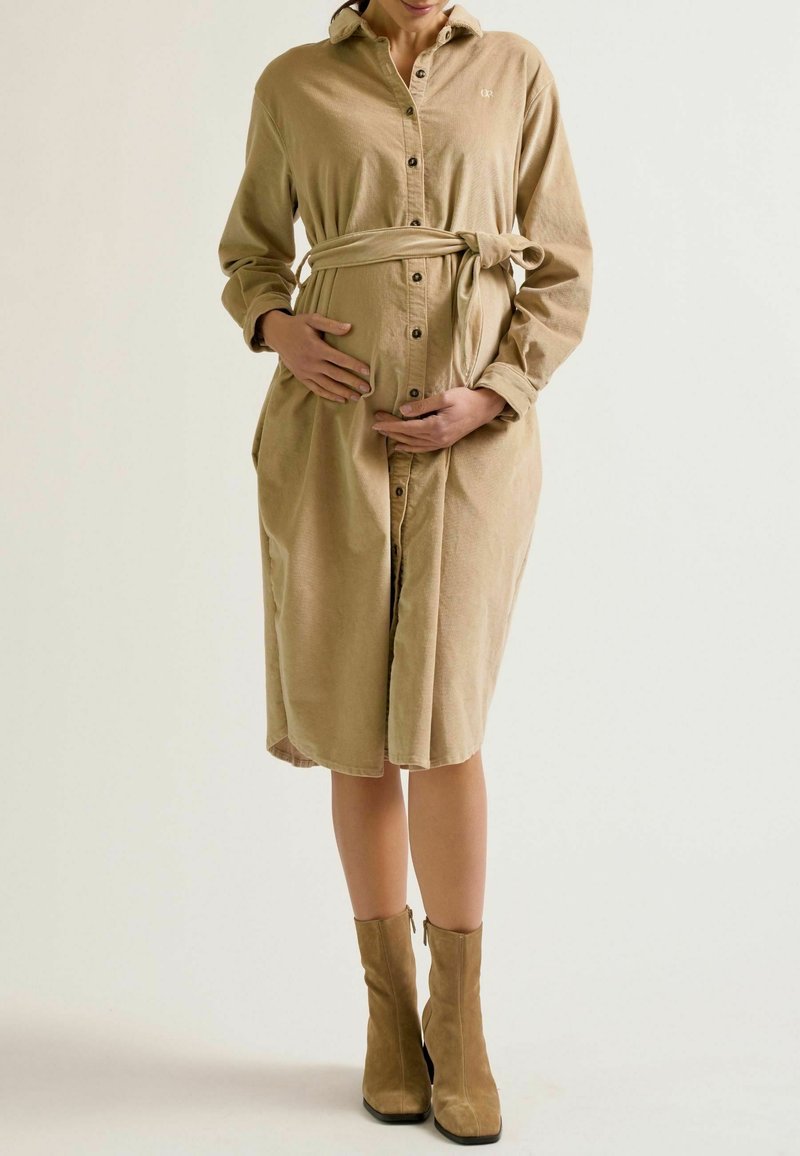 Schwangere Frau in einem beigen Hemdkleid mit gebundenem Taillenzug, die sanft ihren Bauch hält, steht vor einem neutralen Hintergrund.