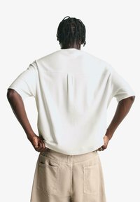 Bílá krátká košile s volným střihem, se zadním yoke a jemným záhybem. V kombinaci s béžovými cargo kalhotami se zadními kapsami.