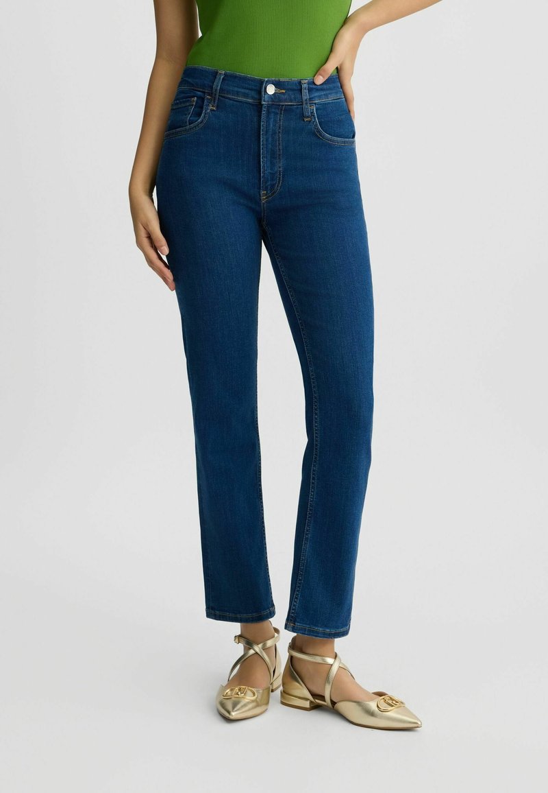 Tmavě modré džíny s vysokým pasem a slim-fit střihem, pět kapes a kontrastní prošití. V kombinaci s plochými botami v metalické zlaté barvě.