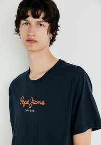 Tamsiai mėlynas medvilninis marškinėliai su oranžiniu logotipo tekstu "Pepe Jeans LONDON" ir klasikinio apvalaus kaklo dizainu. Minkšta medžiaga, trumpomis rankovėmis.