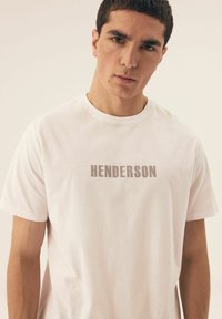 Biała bawełniana koszulka z okrągłym dekoltem i krótkimi rękawami, z dużym szarym napisem "HENDERSON" nadrukowanym na przodzie.