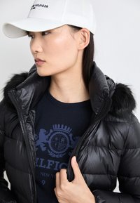 Giacca a puffo nera con finitura lucida, dotata di cappuccio foderato in pelliccia. Indossata sopra una maglietta navy con un design grafico. Berretto da baseball bianco.