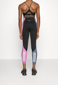 Leggings neri con pannelli laterali rosa e blu, caratterizzati da tre strisce bianche su ciascun polpaccio. Il materiale sembra elastico e aderente.