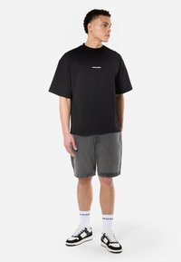 Black short-sleeve t-shirt with white "PEGADOR" text, paired with gray denim shorts and black-and-white sneakers. Plain background.