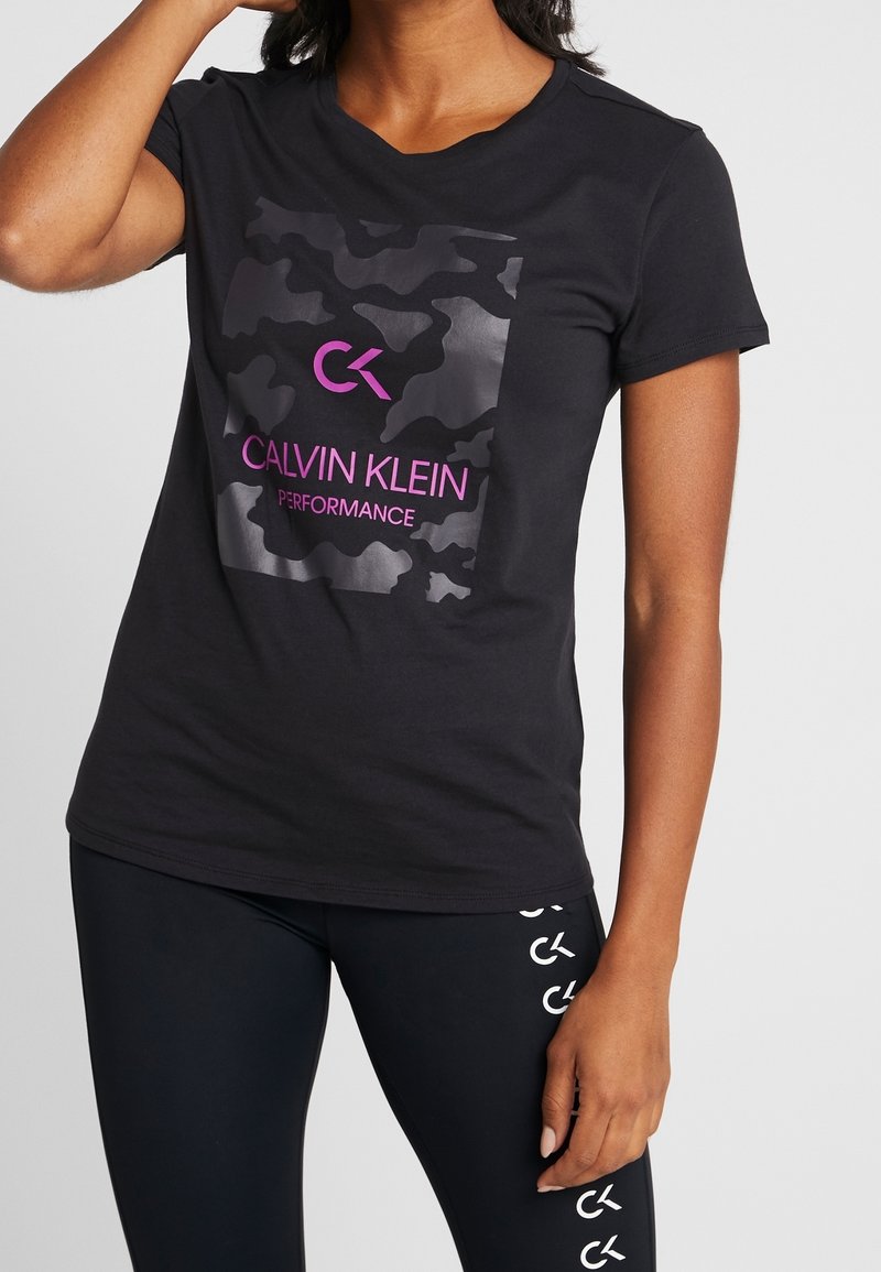 T-shirt en coton noir à manches courtes, présentant un motif camouflage et un logo mis en valeur en rose. Porté avec un legging noir affichant un logo.