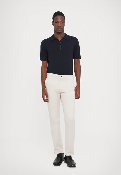 Tricou polo de culoare bleumarin închis, cu fermoar scurt și guler, asortat cu pantaloni bej deschis și pantofi negri. Design simplu, ajustat, cu o textură netedă.