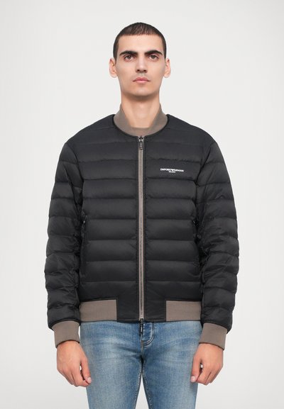 Tânăr îmbrăcat cu o jachetă căptușită Emporio Armani neagră, cu manșete și guler roz pal, asortată cu blugi albaștri deschiși, pe un fundal alb.
