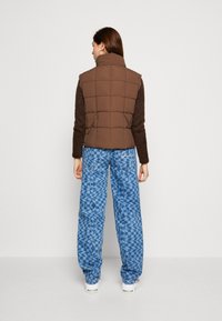 Gilet puffoso marrone con design trapuntato, abbinato a pantaloni blu a fantasia. Il gilet presenta un colletto alto e un tessuto isolante.
