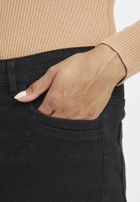 Fekete, slim fit fekvésű farmernadrág elülső zsebbel. Egy kéz a zsebben van. A felső darab bordázott, bézs színű anyagból készült.