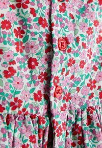 Detailný záber na kvetinovú látku s červenými a ružovými kvetmi, zelenými listami, červenými gombíkmi a záhybami na švíku.