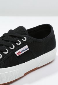 Zapatilla de lona negra con suela de goma blanca, ojales de metal y una etiqueta de marca roja y blanca en el lateral. Diseño clásico de caña baja.
