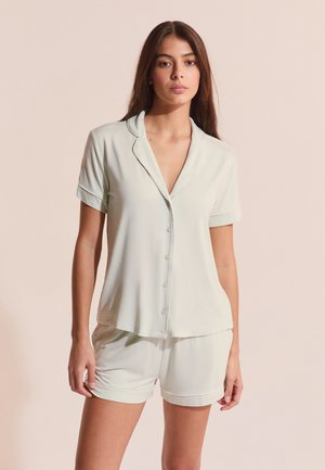 Jeune femme portant une chemise à manches courtes claire boutonnée et un short assorti, posant devant un fond neutre.