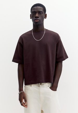 Junger Mann trägt ein lockeres dunkelbraunes T-Shirt, weiße Hose, eine Hand in der Tasche, silberne Kettenhalskette, Armband und Ring, vor einem schlichten Hintergrund.