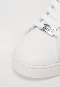 Gros plan sur le bout d’une basket blanche avec des lacets blancs passés dans une pièce métallique argentée gravée du logo "MK".