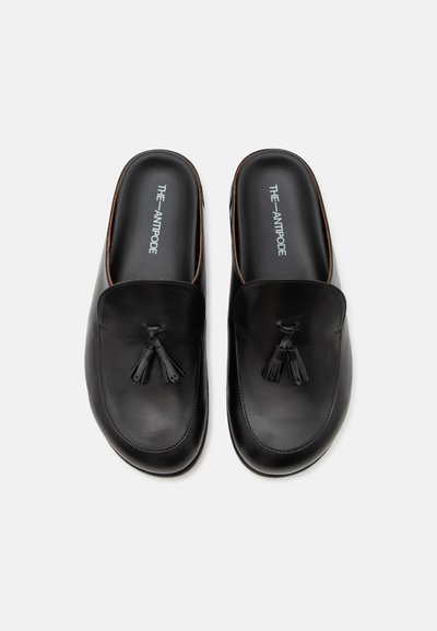 Ζευγάρι μαύρα δερμάτινα loafers χωρίς κορδόνια με φούντες, εμφανές το όνομα της μάρκας "THE ANTIPODE" μέσα σε κάθε παπούτσι.