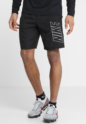 Mężczyzna w czarnych szortach Nike z dużym logo na prawej nodze, czarnych skarpetkach Nike oraz białych sneakersach Nike z czerwonym znakiem.