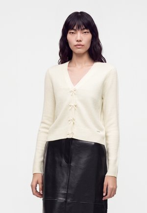 Vrouw met lang zwart haar die een crèmekleurige cardigan met strikjes draagt en een zwarte leren rok tegen een effen witte achtergrond.