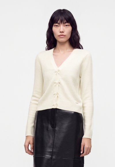 Femme aux longs cheveux noirs portant un cardigan crème avec des nœuds papillon et une jupe en cuir noire sur fond blanc uni.