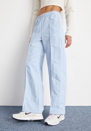 Personne portant un pantalon Nike large bleu clair, un pull court blanc et des baskets Nike blanches, debout sur un sol moquetté bleu.