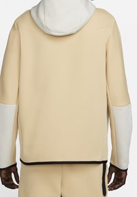 Beżowa bluza z kapturem, z białym kapturem i białymi łatami na łokciach, noszona przez osobę pokazana z tyłu, z czarnym wykończeniem na mankietach i u dołu.