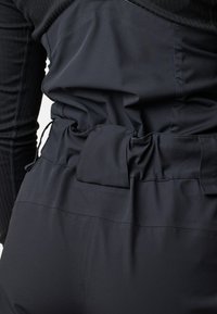 Lähikuva mustasta kangasvaatteesta, jossa on kerätty vyötärö leveällä vyönkaltaisella yksityiskohdalla ja vasemmassa kädessä oleva ribbineulottu pitkä hiha.