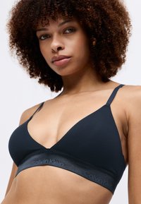 Soutien-gorge triangle noir en tissu lisse, avec de fines bretelles ajustables et une bande élastique de marque portant "Calvin Klein" en gris.