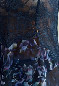 Top azul marino de encaje con parte inferior de gasa con estampado floral, que presenta un dobladillo de encaje festoneado y un detalle de logo dorado cerca de la abertura, con una textura delicada.