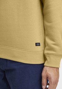 Yellow sweatshirt with ribbed cuffs and hem, featuring a small black label on the lower left side. Smooth texture and relaxed fit.