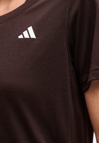 Κοντινό πλάνο ενός ατόμου που φοράει σκούρο καφέ πουκάμισο με λευκό λογότυπο Adidas στο πάνω μέρος του στήθους κοντά στον ώμο.