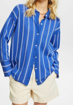 Camisa de botones a rayas azul con cuello y mangas largas. Presenta rayas verticales blancas y botones blancos. Combinada con pantalones cortos claros.