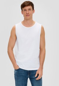 Chemise en coton blanche sans manches avec un col rond, associée à un jean en denim bleu. Le mannequin se tient avec une main dans la poche devant un fond uni.