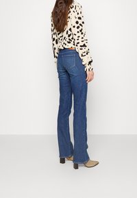 Femme aux longs cheveux bruns portant un chemisier blanc à pois noirs, un jean évasé bleu et des bottines beiges à talons, debout devant un fond blanc.