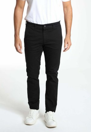 Homme portant un pantalon noir, des baskets blanches et un t-shirt blanc, debout devant un fond blanc uni.