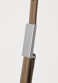 Bracelet en cuir marron avec un fermoir en métal argenté rehaussé d'un logo gravé. Texture lisse avec des détails de couture subtils.