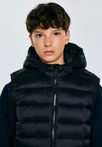Schwarze gepolsterte Weste mit Kapuze, mit einem Reißverschluss vorne, glattem Stoff und gestepptem Design. Getragen über einem dunklen Langarmshirt.