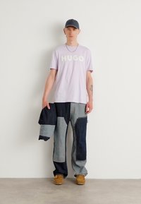 Light purple t-shirt with "HUGO" in bold letters, paired with patchwork denim pants in various shades of blue and brown work boots.