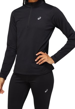 Femme portant un haut de sport noir à manches longues avec une fermeture éclair au quart et des leggings noirs assortis avec des logos Asics.