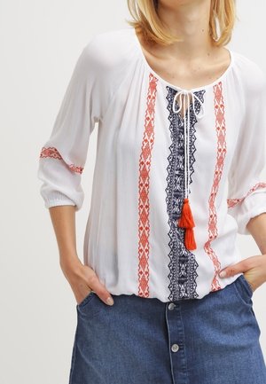 Vrouw die een witte blouse draagt met rode en zwarte borduur, een gestrikte halslijn met tassels en een blauwe denim rok met knopen.