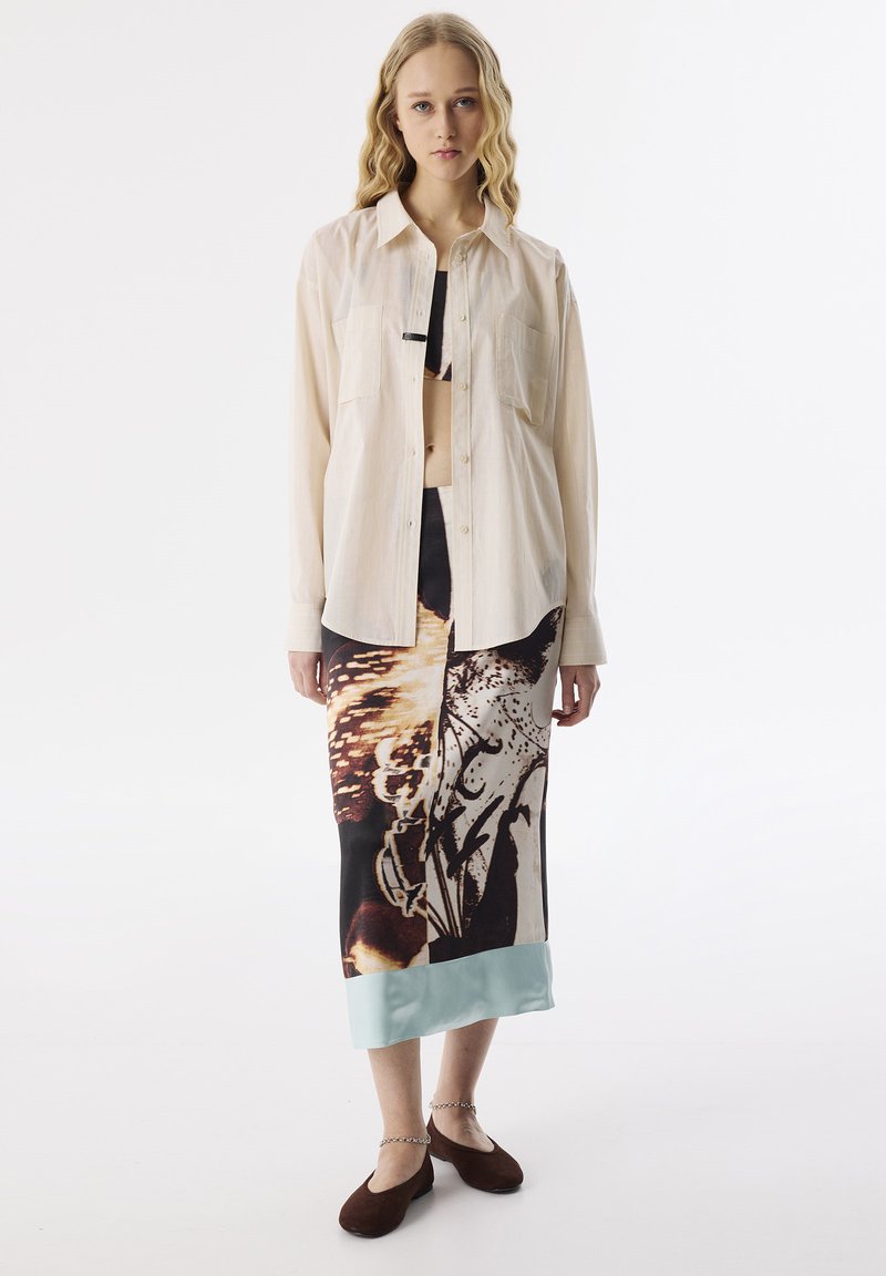 Jasno beżowa koszula z guzikami i dwoma kieszeniami na piersi, w zestawieniu z czarną i beżową spódnicą z nadrukiem, posiadającą jasnoniebieski satynowy dół. Brązowe baleriny.