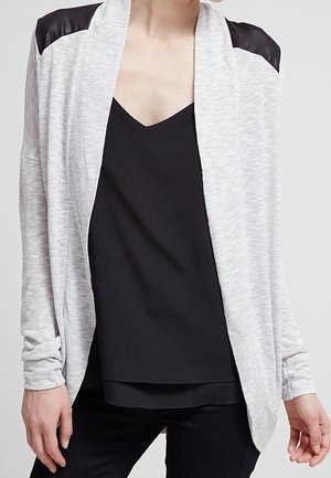 Personne portant un cardigan gris clair ouvert sur le devant avec des empiècements noirs aux épaules, par-dessus un haut noir à col en V et un pantalon noir.
