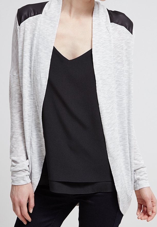 Personne portant un cardigan gris clair ouvert sur le devant avec des empiècements noirs aux épaules, par-dessus un haut noir à col en V et un pantalon noir.