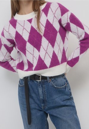 Personne portant un pull à losanges violet et blanc rentré dans un jean bleu taille haute avec une ceinture noire.