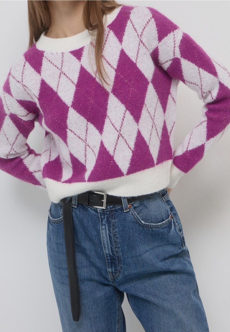 Personne portant un pull à losanges violet et blanc rentré dans un jean bleu taille haute avec une ceinture noire.