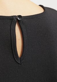 Černý top s malým otvorem na zádech, který je zajištěn černým knoflíkem, vyrobený z texturované látky s viditelným švem.