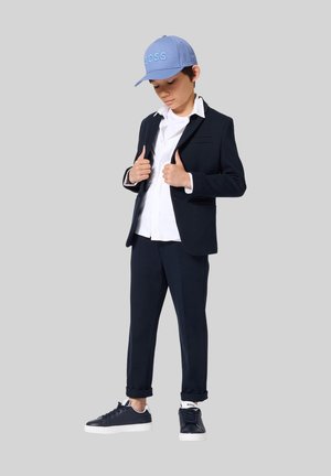 Jongen in een marineblauw pak met opgerolde broekspijpen, wit overhemd, marineblauwe sneakers en een lichtblauwe pet die zijn jasje rechttrekt tegen een effen achtergrond.