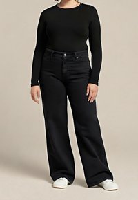 Personne portant un haut noir à manches longues, un pantalon noir taille haute à jambes larges, et des chaussures blanches sur un fond beige uni.