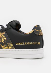 Black and gold Versace Jeans Couture sneaker with white sole, decorative baroque pattern, and white shoelaces, side view detail.