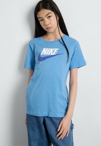 Světle modré bavlněné tričko s krátkými rukávy, na kterém je výrazné bílé a fialové logo Nike napříč hrudí. Jednoduchý kulatý výstřih.